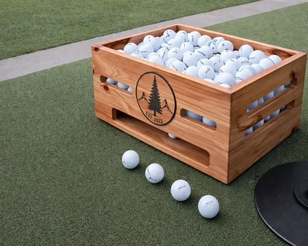 Custom wooden range ball basket with engraved club logo, filled with golf balls on a driving range.