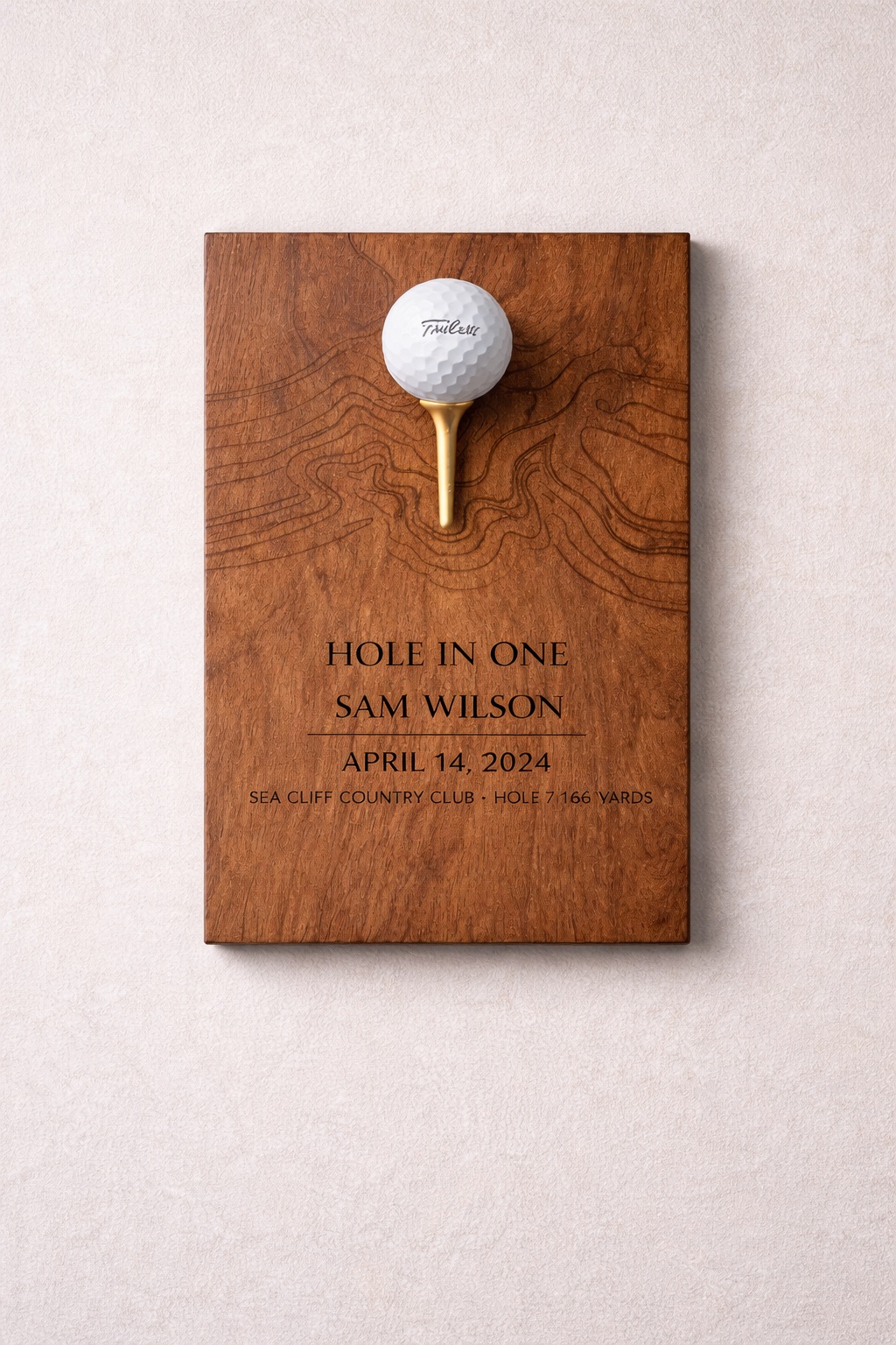 Close-up of a walnut hole-in-one plaque with engraved topographic contour lines, golf ball on gold tee, light background.