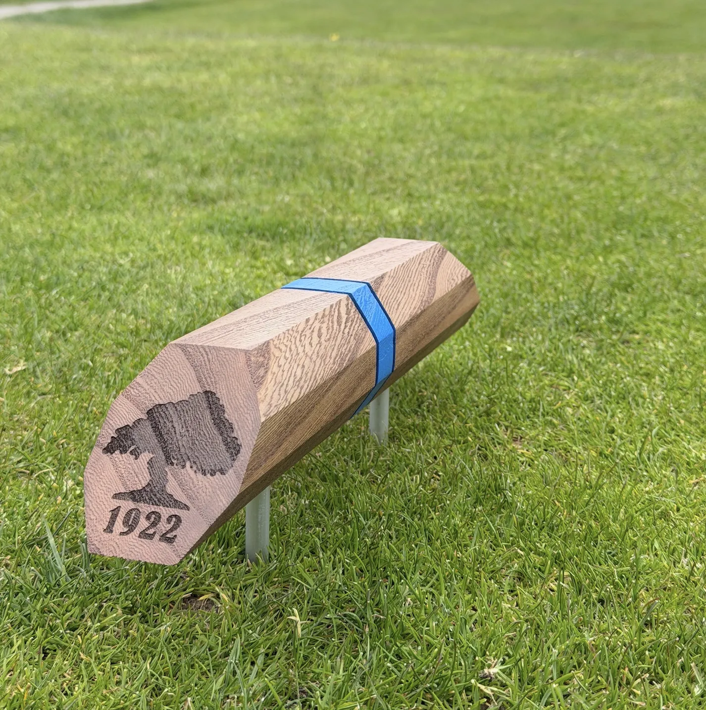 A walnut hexagonal tee marker with blue inlay stripe on steel legs, sitting on turf.